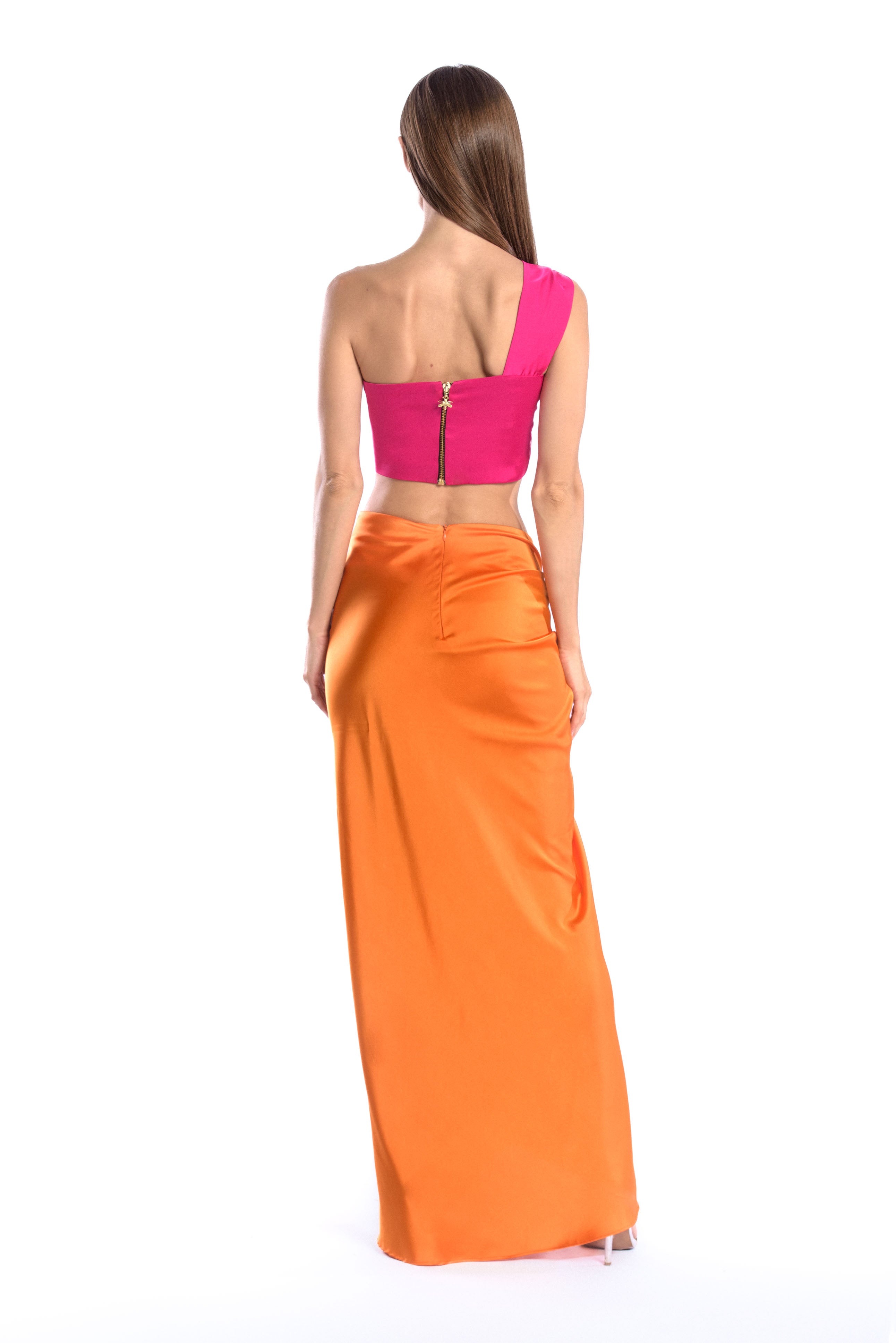 Back shot of model wearing a silk cross shoulder crop top in hot pink or fuchsia with back firefly logo golden zipper and silk skirt in orange color with a seashell brooch