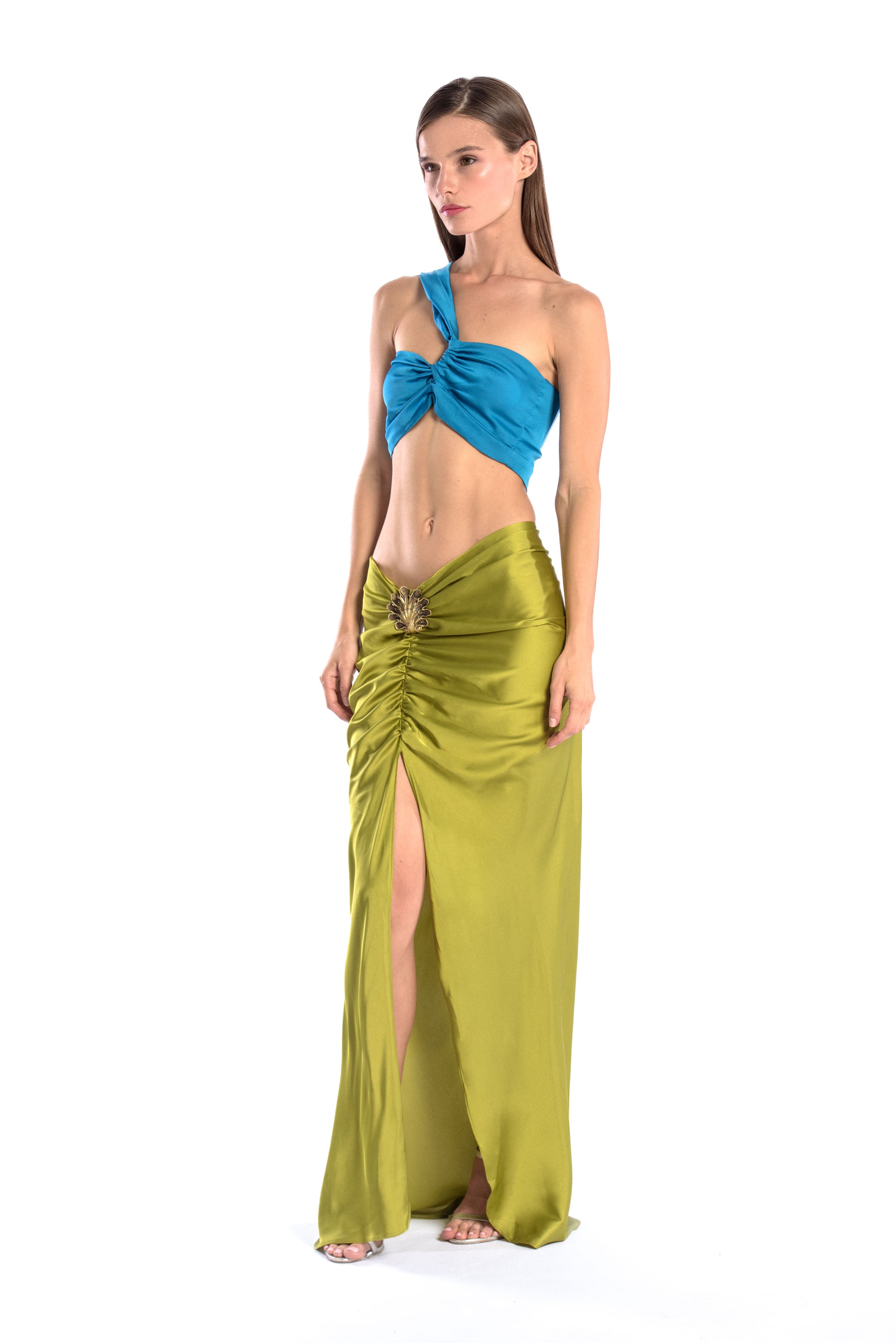 Model wearing a silk cross shoulder crop top in blue and silk skirt in olive color with a seashell brooch
