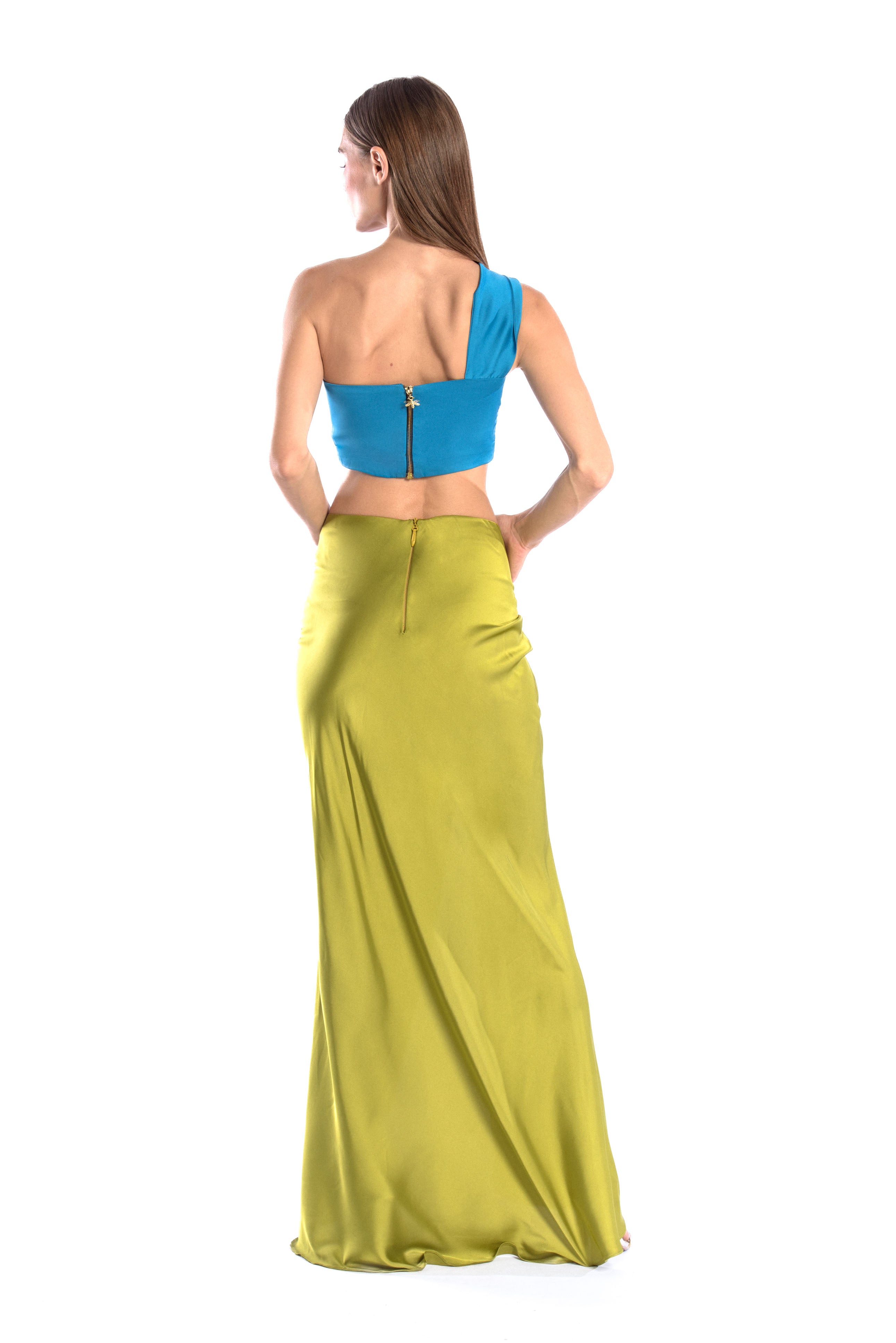 Back shot of model wearing a silk cross shoulder crop top in blue with back firefly logo golden zipper and silk skirt in olive color with a seashell brooch