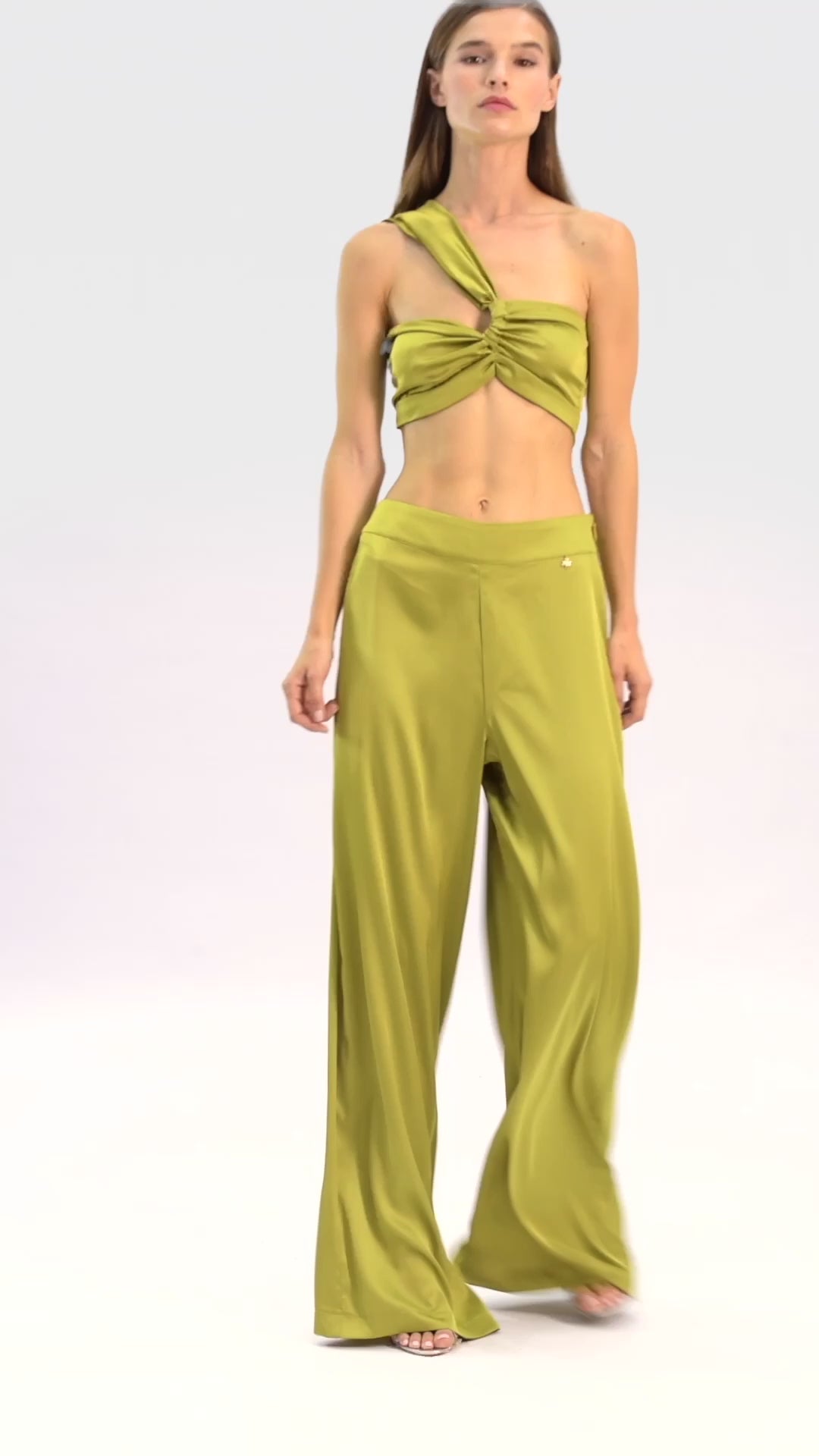 Video of model wearing a silk cross shoulder crop top in olive with back firefly logo golden zipper and silk pants in olive with a firefly golden charm on white background