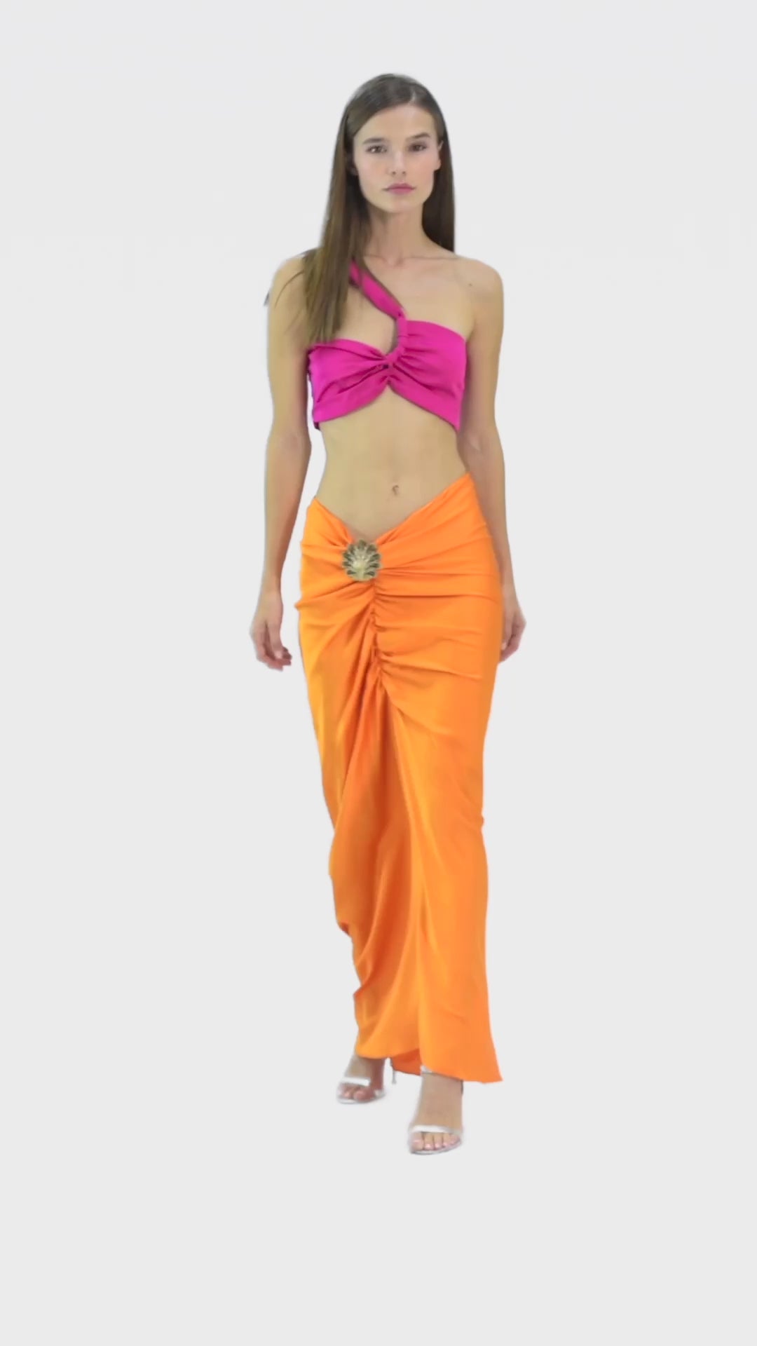 Model wearing a silk cross shoulder crop top in hot pink or fuchsia with back firefly logo golden zipper and silk skirt in orange color with a seashell brooch